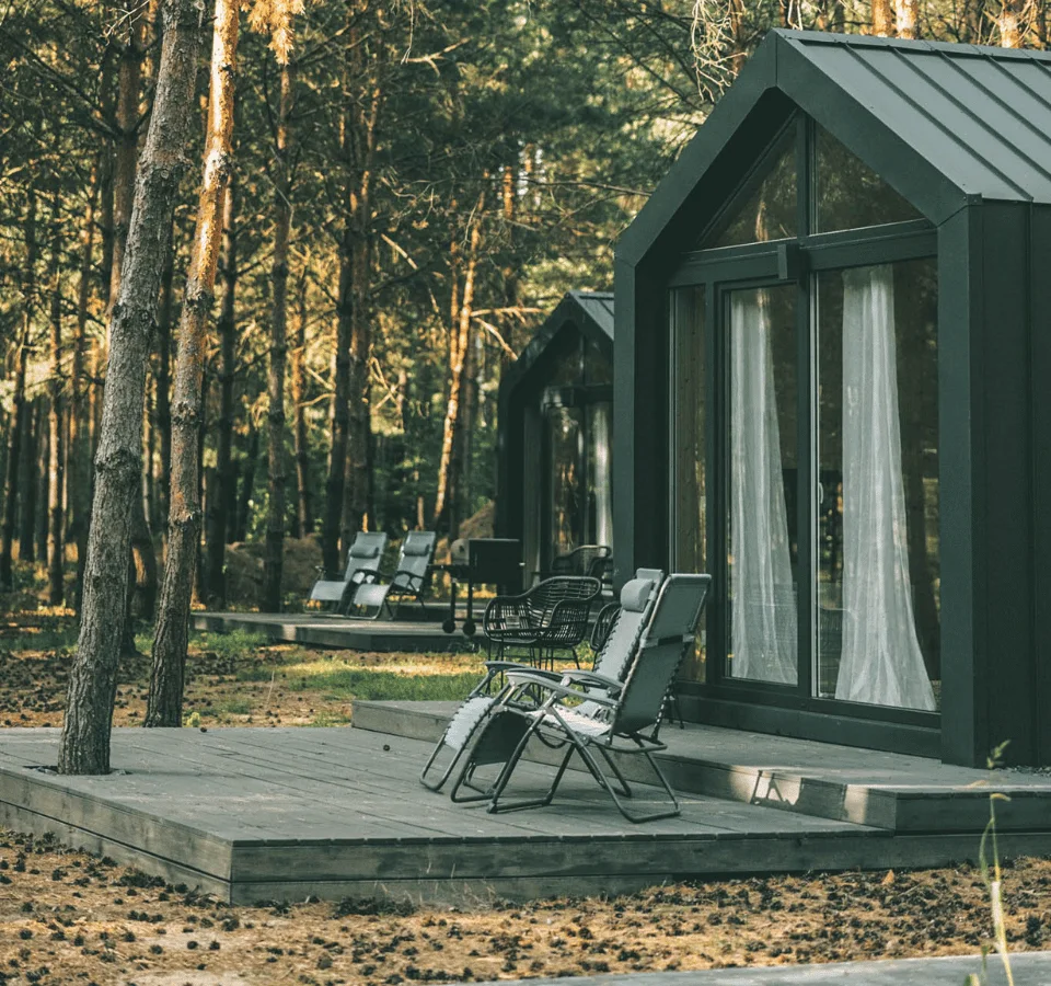 Tiny house with chairs and a wooden terrace, large windows, surrounded by trees, and brown soil.
