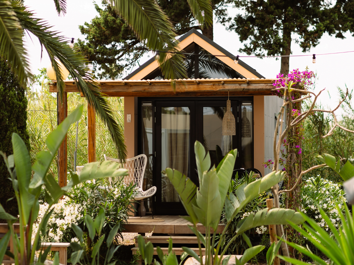 Tiny house surrounded by trees and bushes, wooden terrace, steel frame with wooden finish, palm tree, Ibiza, Spain